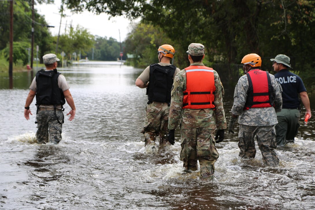 A group of soldiers and a police officer walk through knee deep water in a flooded street.