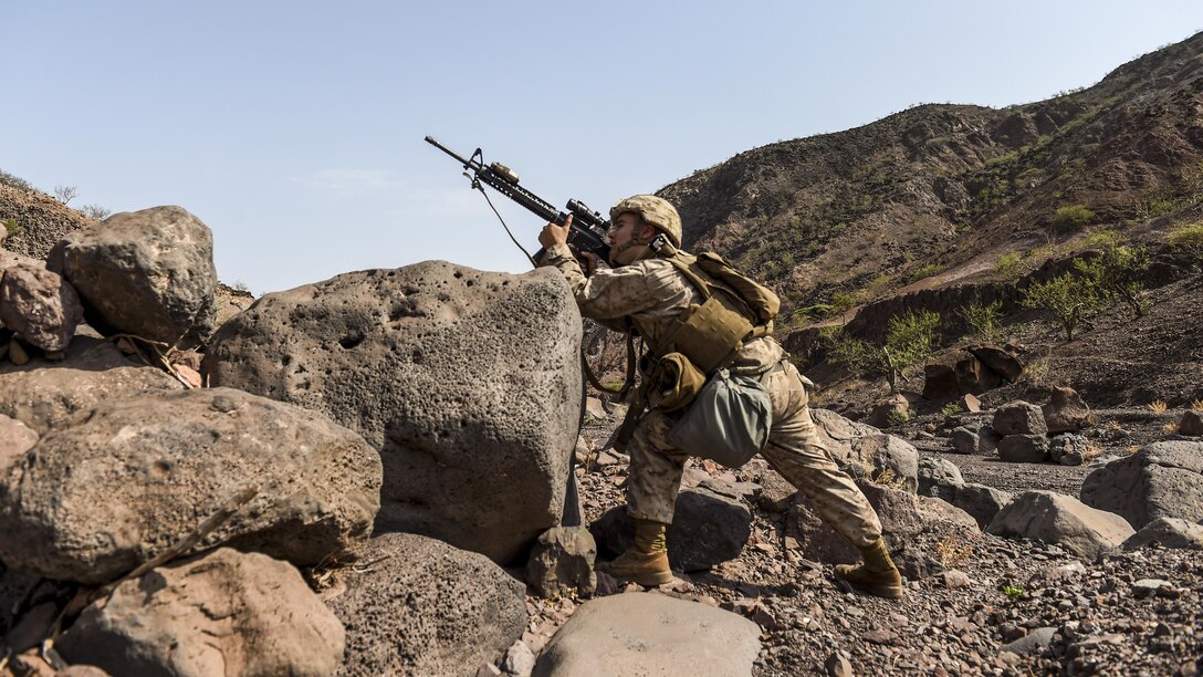 A Marine aims his rifle up a rocky hill.