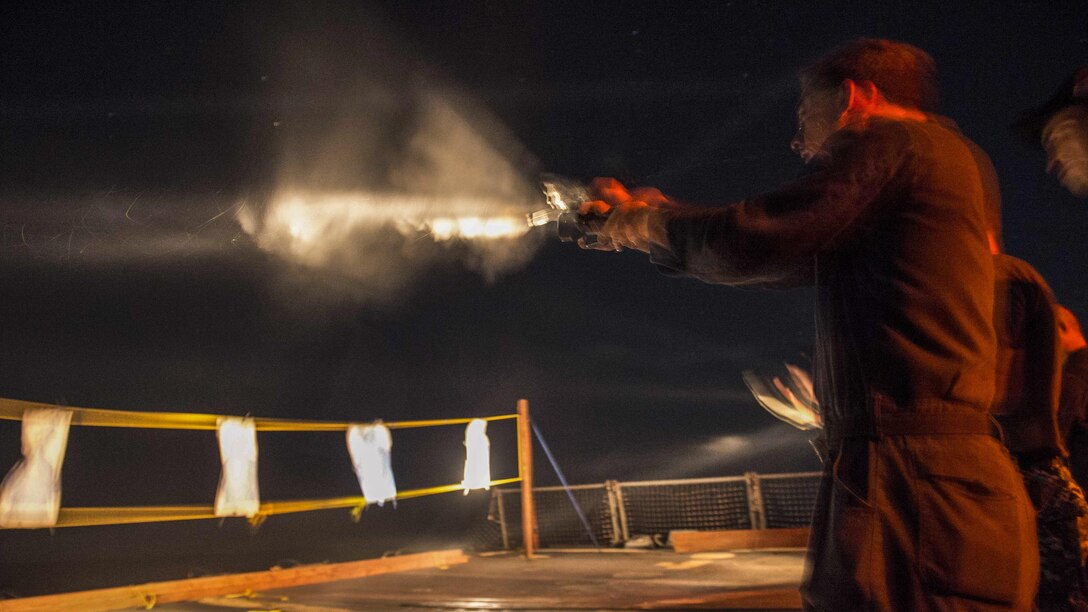 A man fires a handgun at night from a ship.