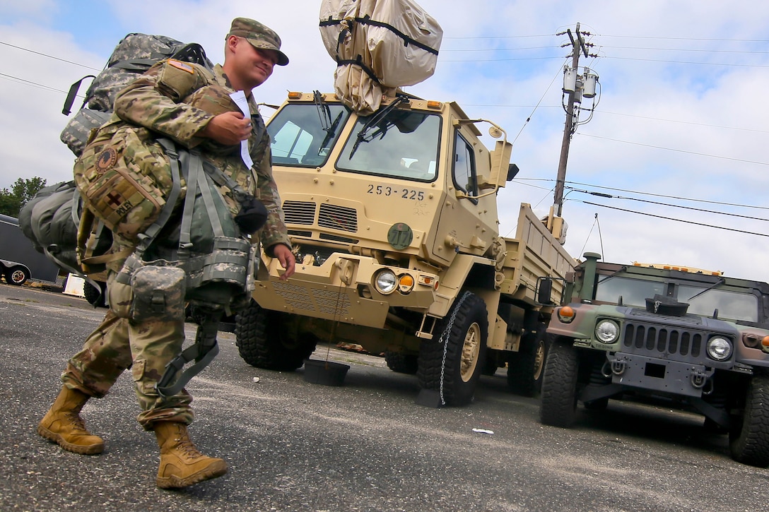 A soldier carrying bags and equipment walks past military vehicles.