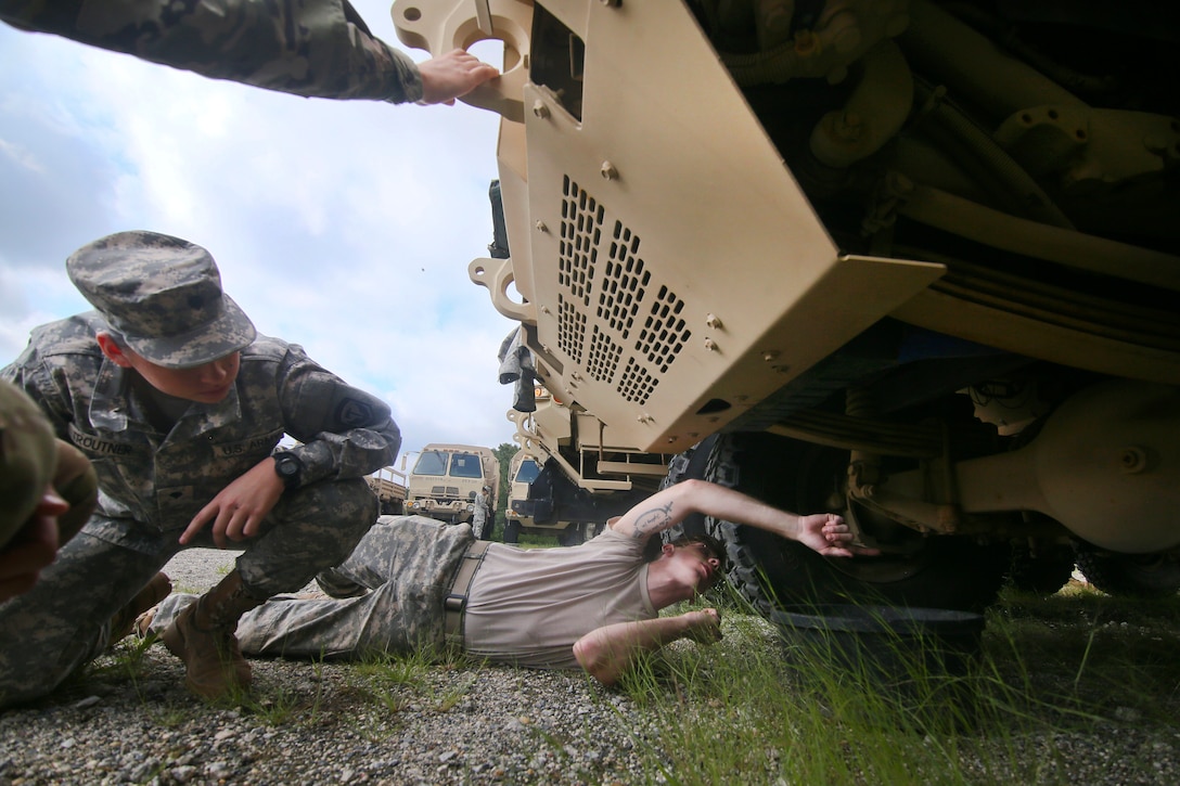 A soldier lays on the ground while another kneels to look underneath a military vehicle.