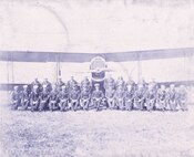 IMAGE: Group Photo of 27 aviation personnel in two rows in front of a single propeller, b plane.