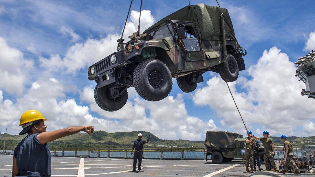 Troops on a flight deck lower a Humvee suspended by metal cords.