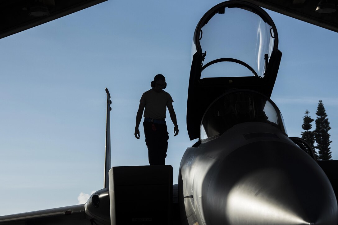 An airman, shown in silhouette, walks on an aircraft with its cockpit hatch open.
