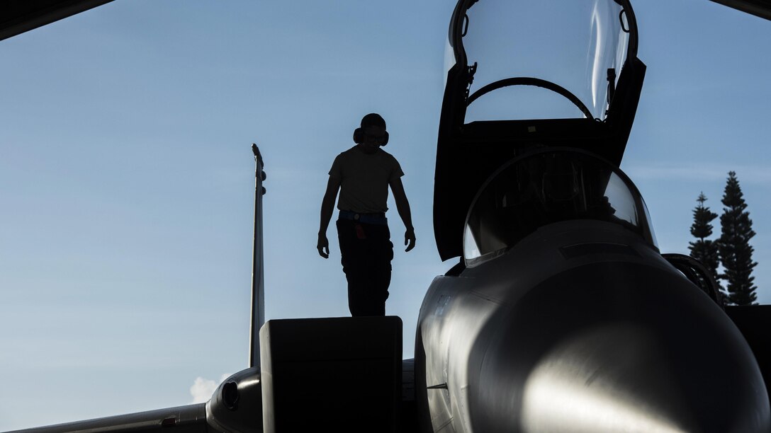 An airman, shown in silhouette, walks on an aircraft with its cockpit hatch open.