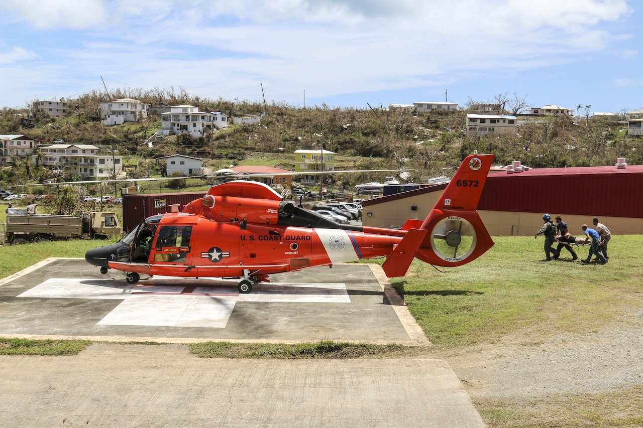 Service members carry a stretcher toward an orange helicopter on a landing pad.