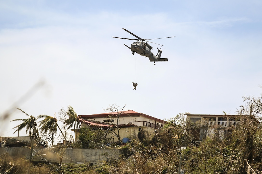 Service members hang on a line from a helicopter hovering over damaged tropical landscape.