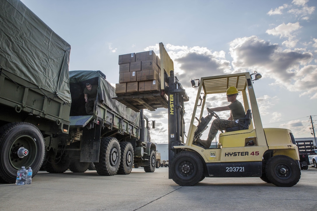 A Marine uses a forklift to lift cardboard boxes near a waiting truck.