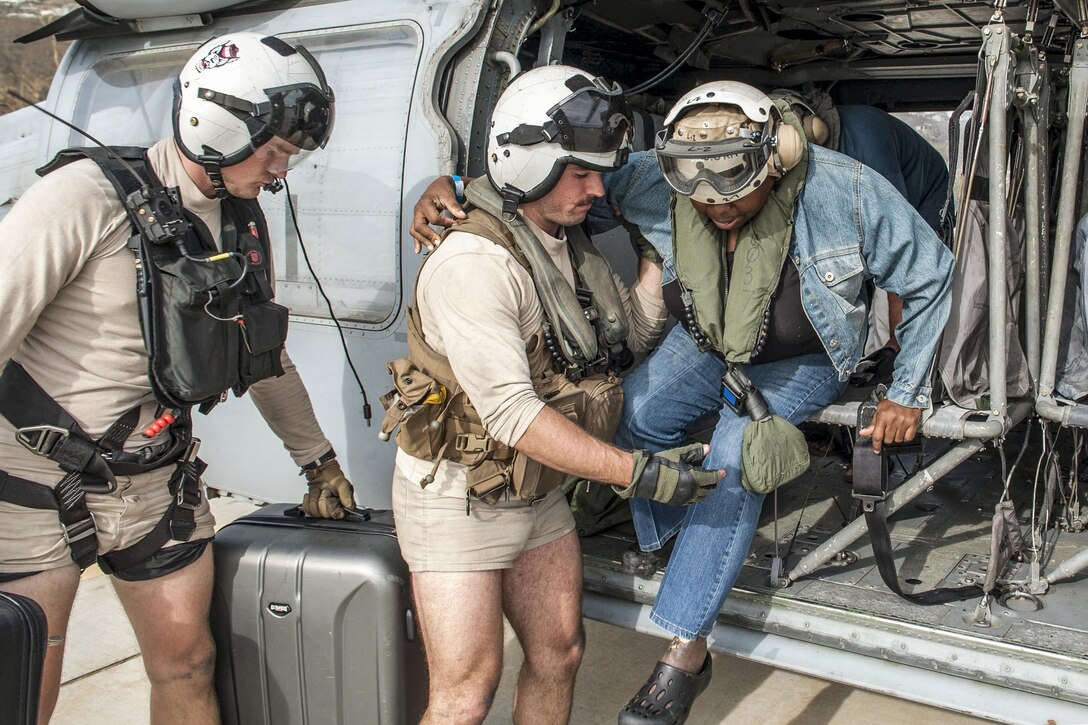 A sailor assists a citizen in a helicopter, as another sailor stands by with a suitcase.