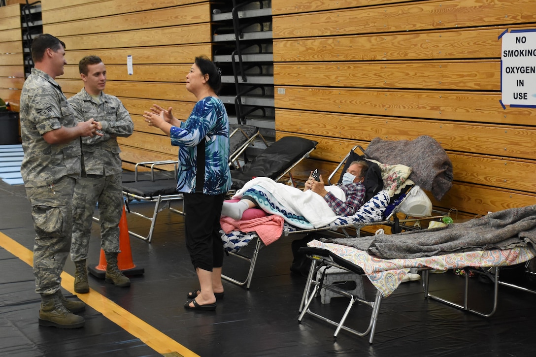 A woman speaks to two military personnel while a man wearing a face mask lays on a cot.