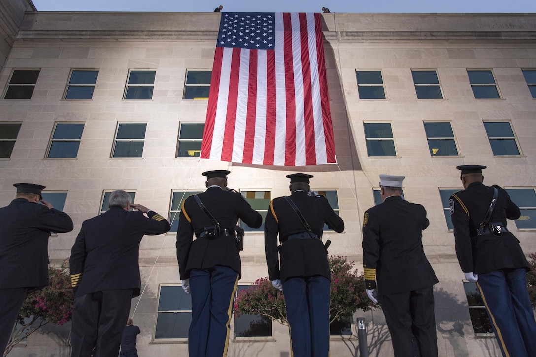 Service members look up to salute an American flag that hands off the roof of the Pentagon.