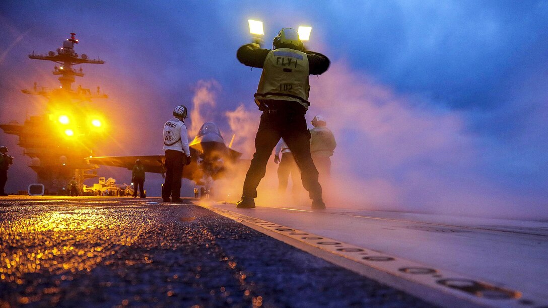 Sailors wave an aircraft onto a runway with yellow lights in the background.