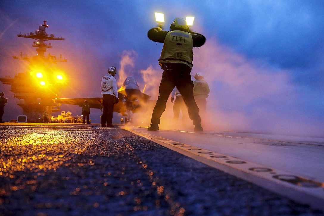 A sailor waves an aircraft onto a runway with yellow lights in the background.