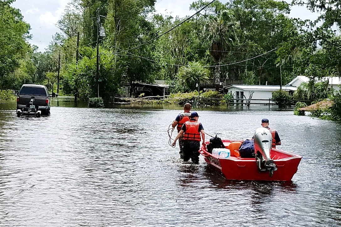 Members of the Coast Guard walk in kneed deep flood waters pulling a boat.