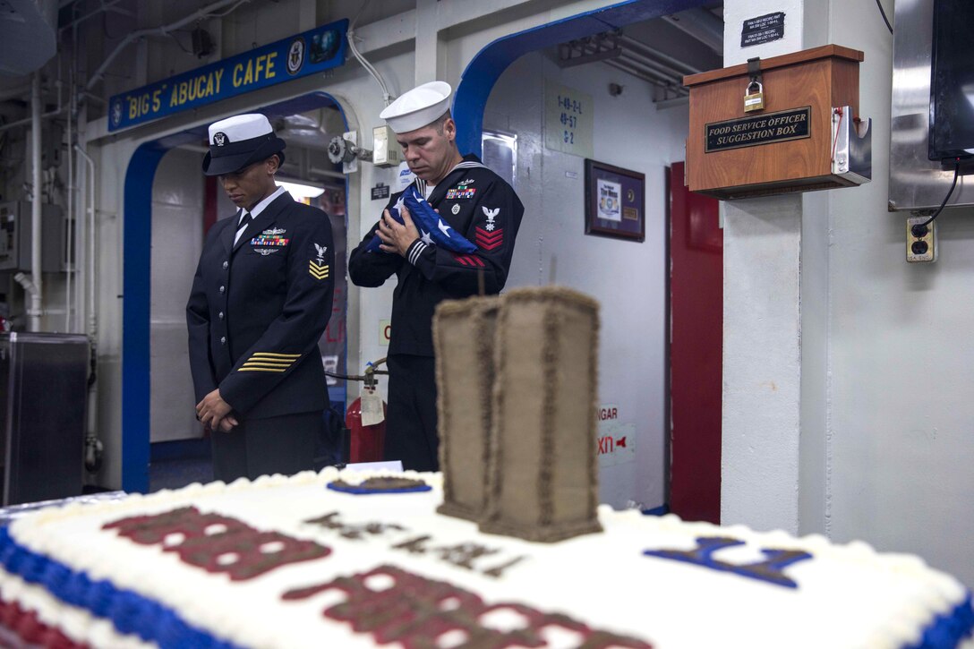 Two sailors stand, one holding a folded flag.