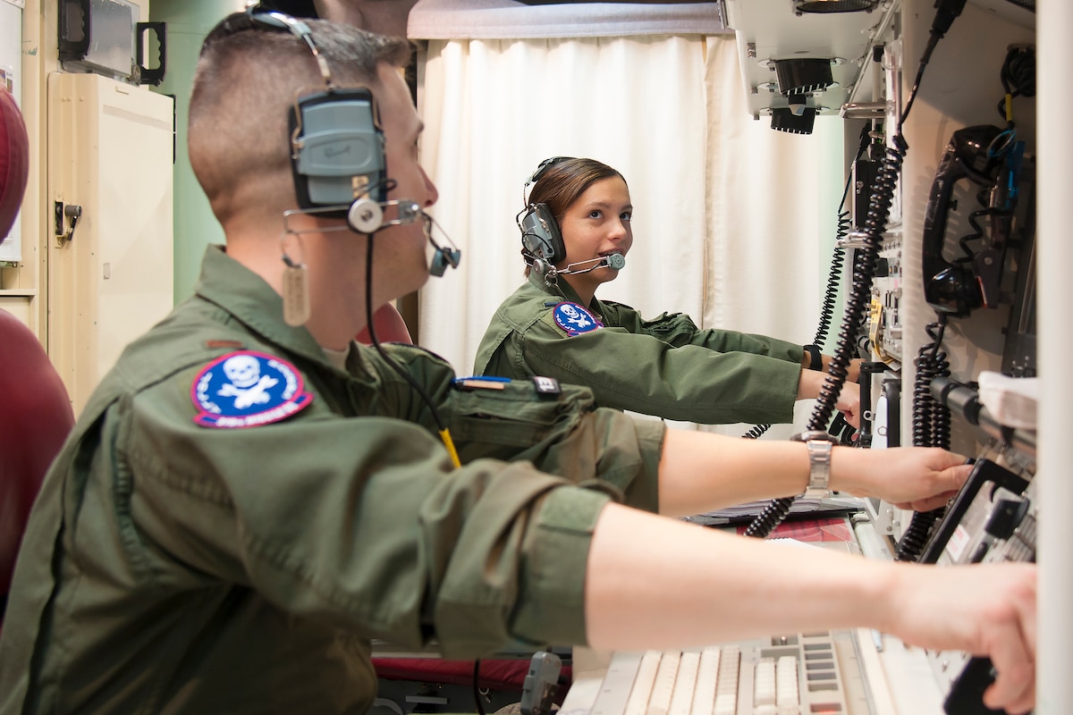 Two airmen have their hands on controls while sitting at a control panel wearing headsets.