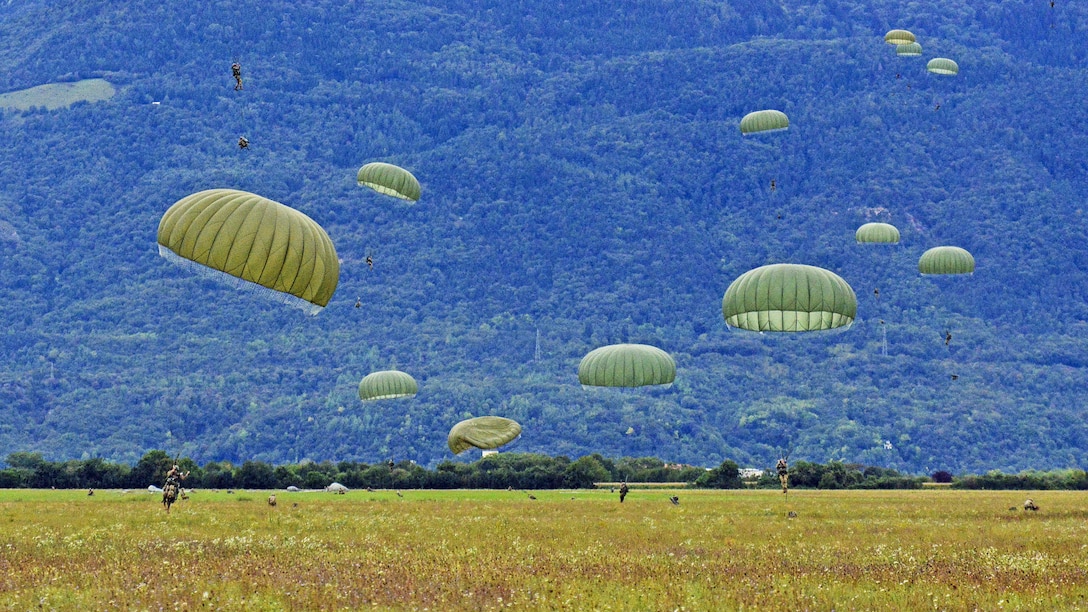 Soldiers with green parachutes land on green grass in front of a blue hill.