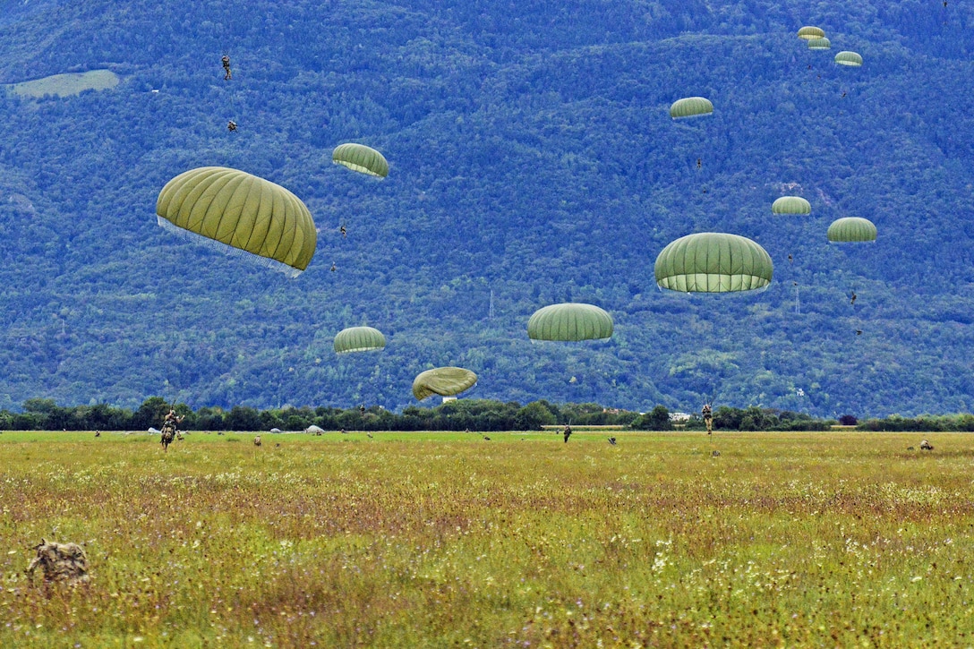 Soldiers with green parachutes land on green grass in front of a blue hill.
