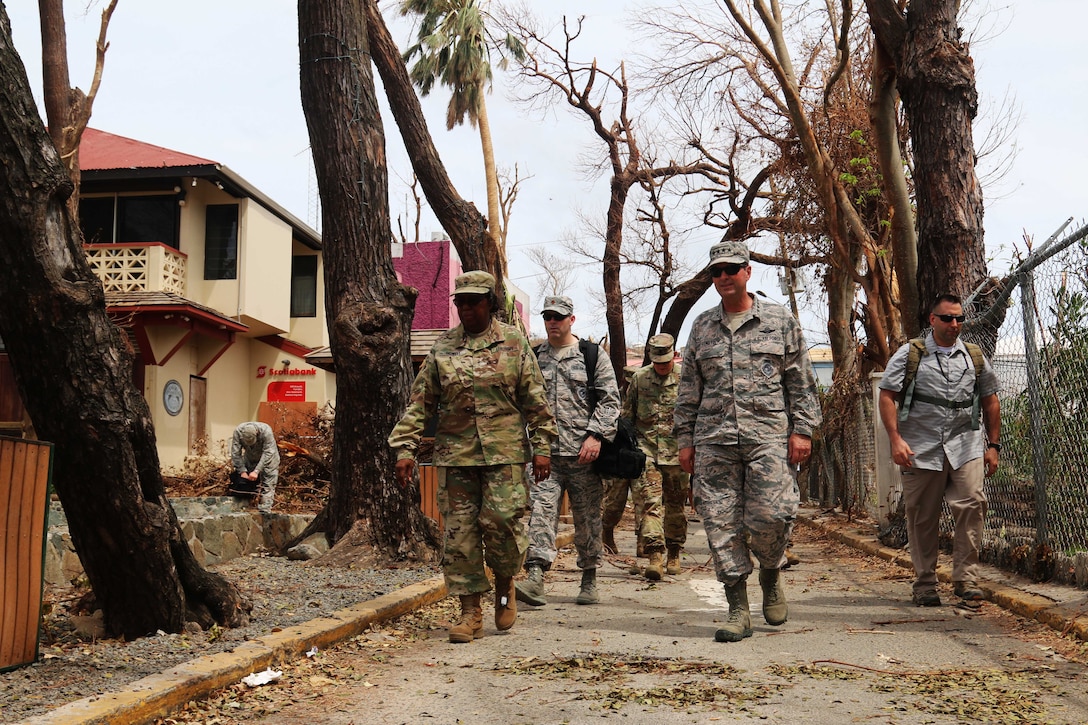 A group of service members and civilians walk down a road with damage to trees and structures.