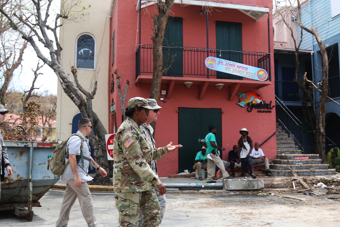 Service members and civilians walk down a street with damage to trees and buildings.