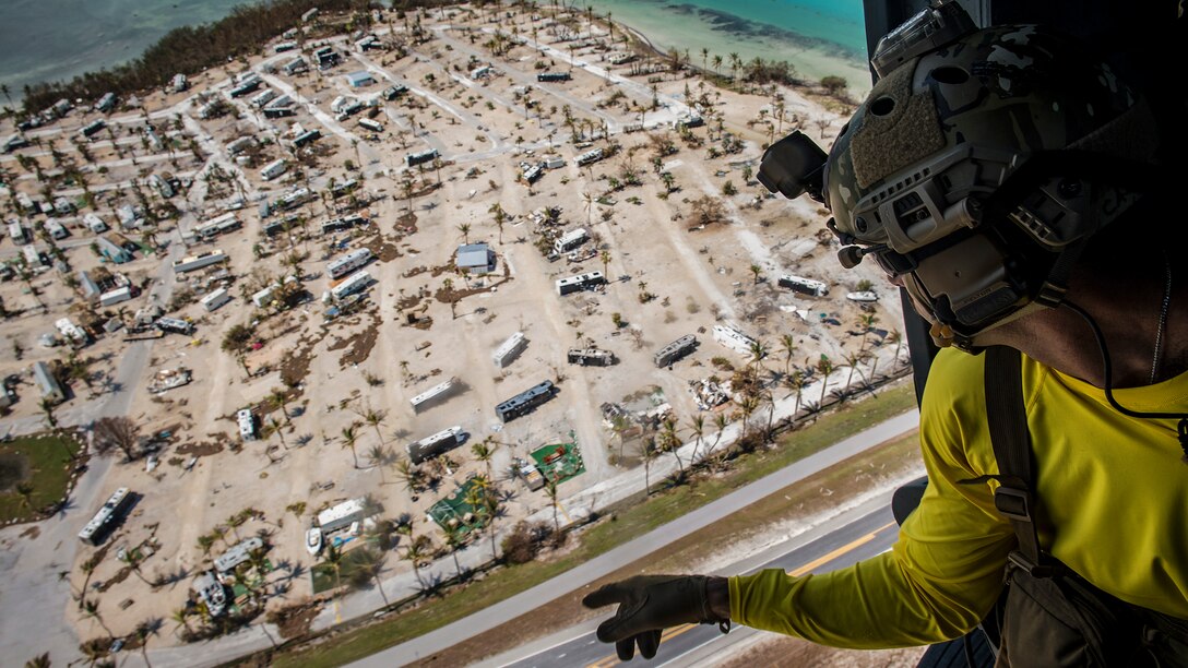 An airman aboard a helicopter points while looking over a damaged beachfront trailer park.