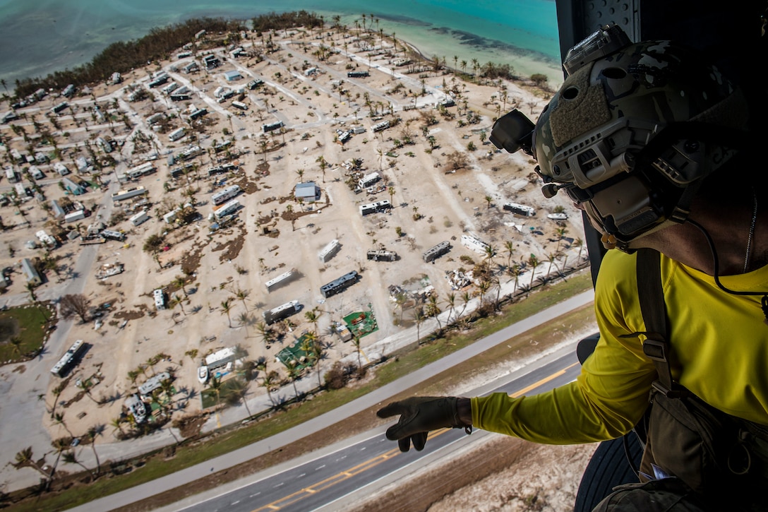 An airman aboard a helicopter points while looking over a damaged beachfront trailer park.