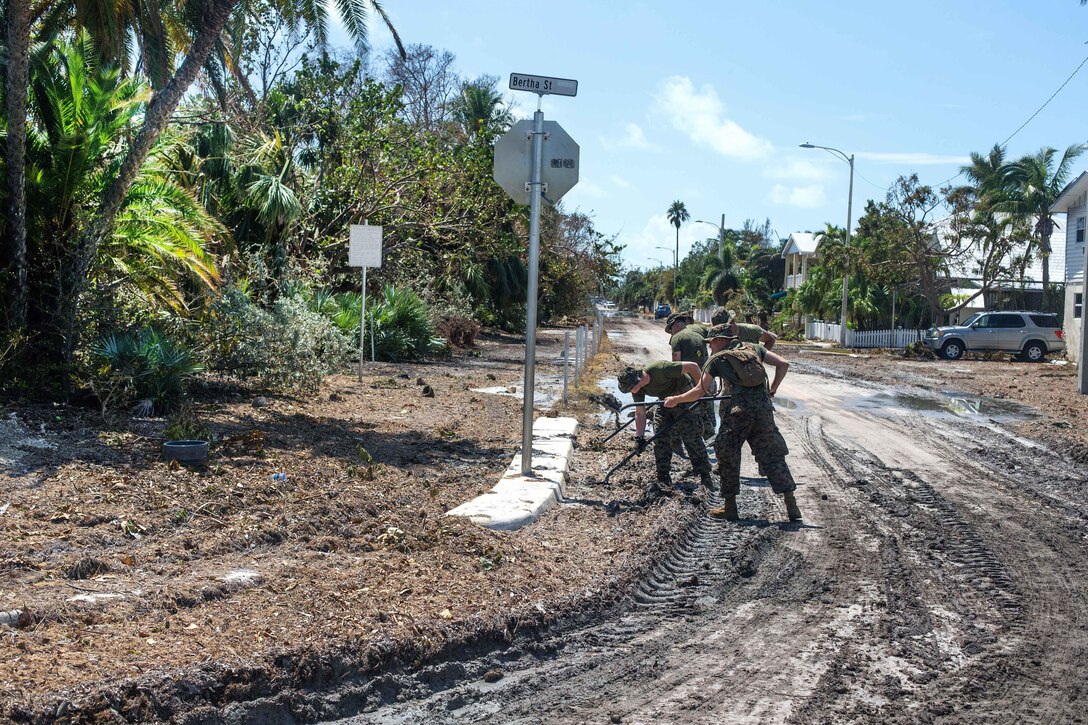 A row of Marines use shovels to clear debris from the street.