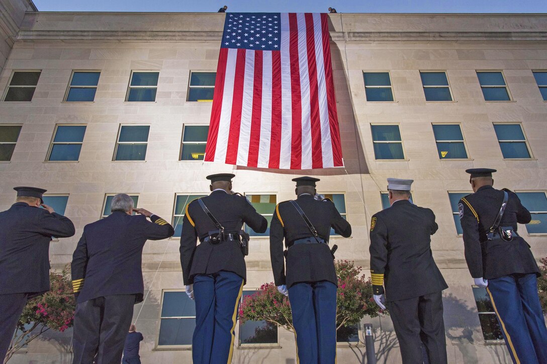 Service members look up to salute an American flag that hands off the roof of the Pentagon.