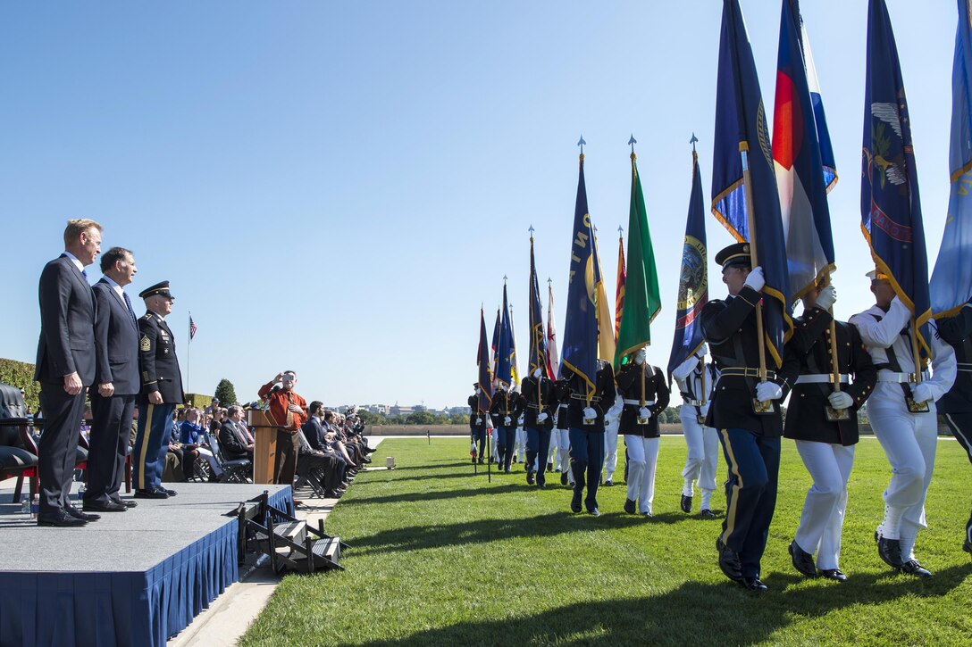 Three Defense Department leaders watch service members march with flags.