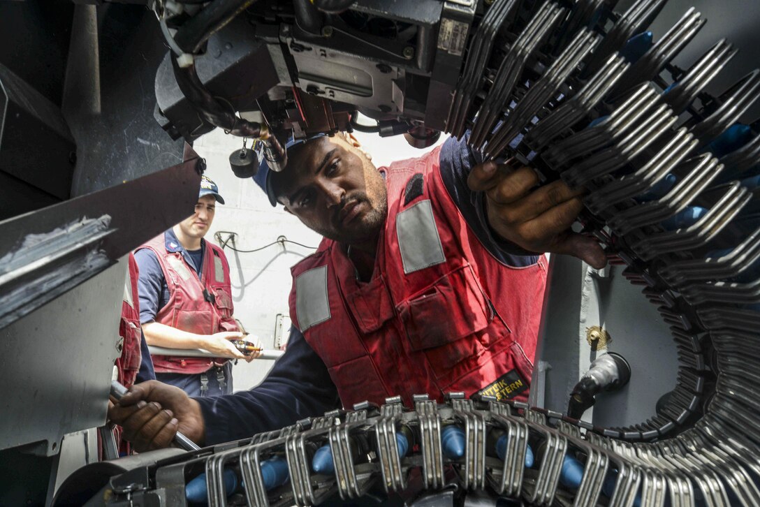 A sailor is framed by a machine gun belt as he loads ammunition.