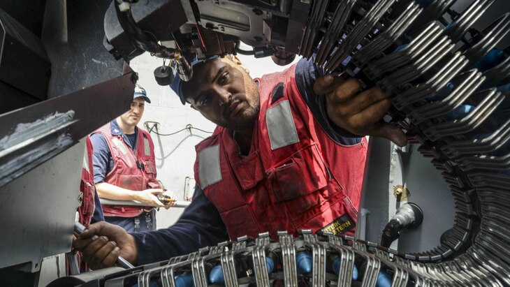 A sailor is framed by a machine gun belt as he loads ammunition.