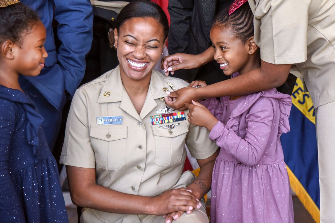A woman kneels and smiles while a girl helps pin anchors to her uniform collar.