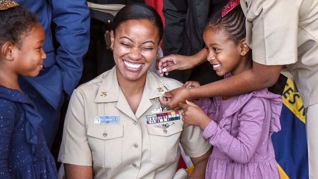 A woman kneels and smiles while a girl helps pin anchors to her uniform collar.
