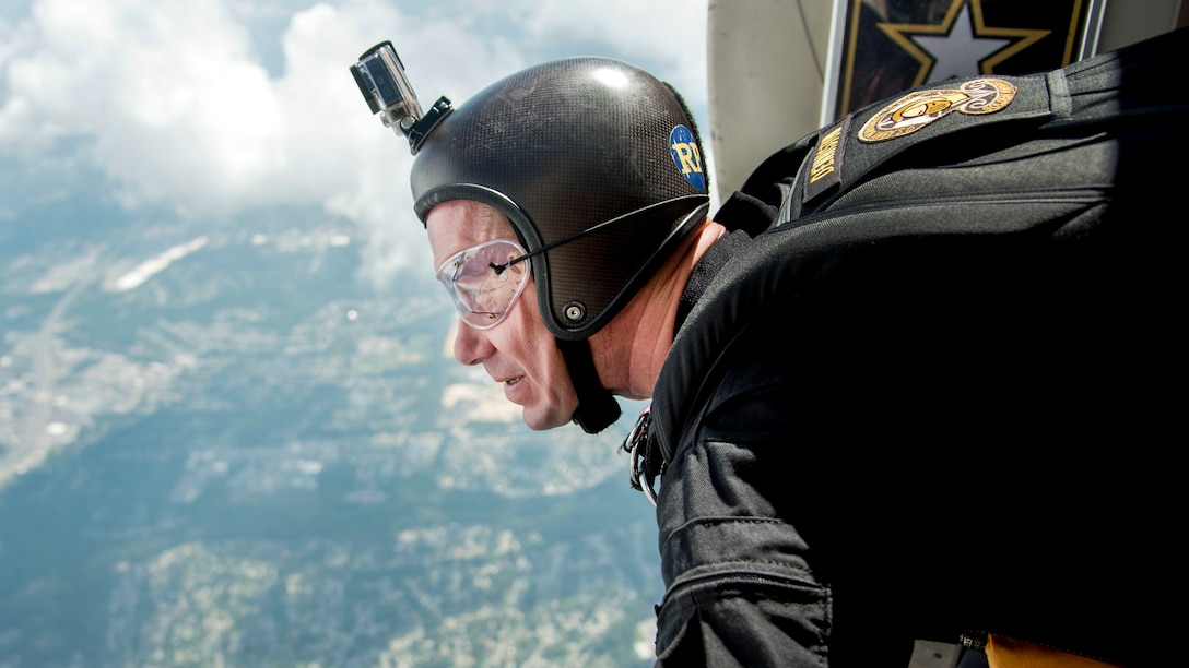 A soldier wearing a helmet and goggles looks out from an open aircraft door.