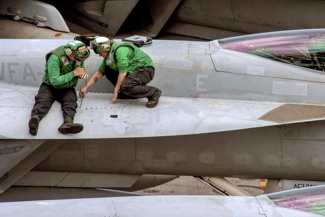 Two sailors in green shirts work on the wing of an aircraft.