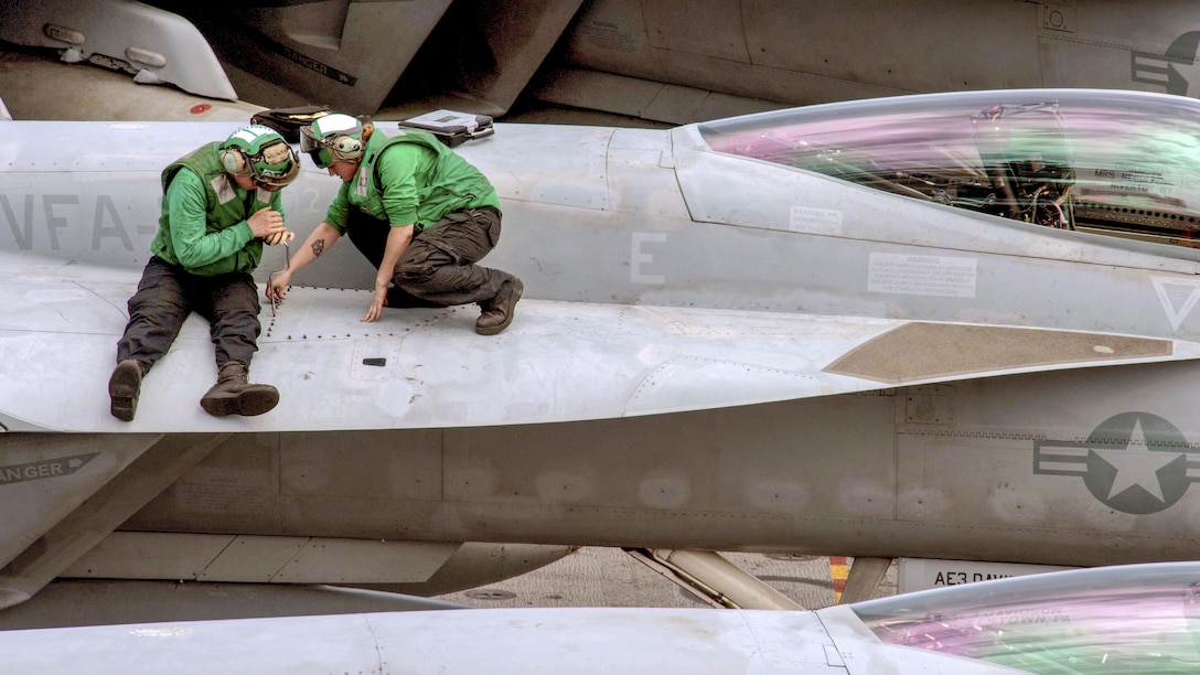 Two sailors in green shirts work on the wing of an aircraft.