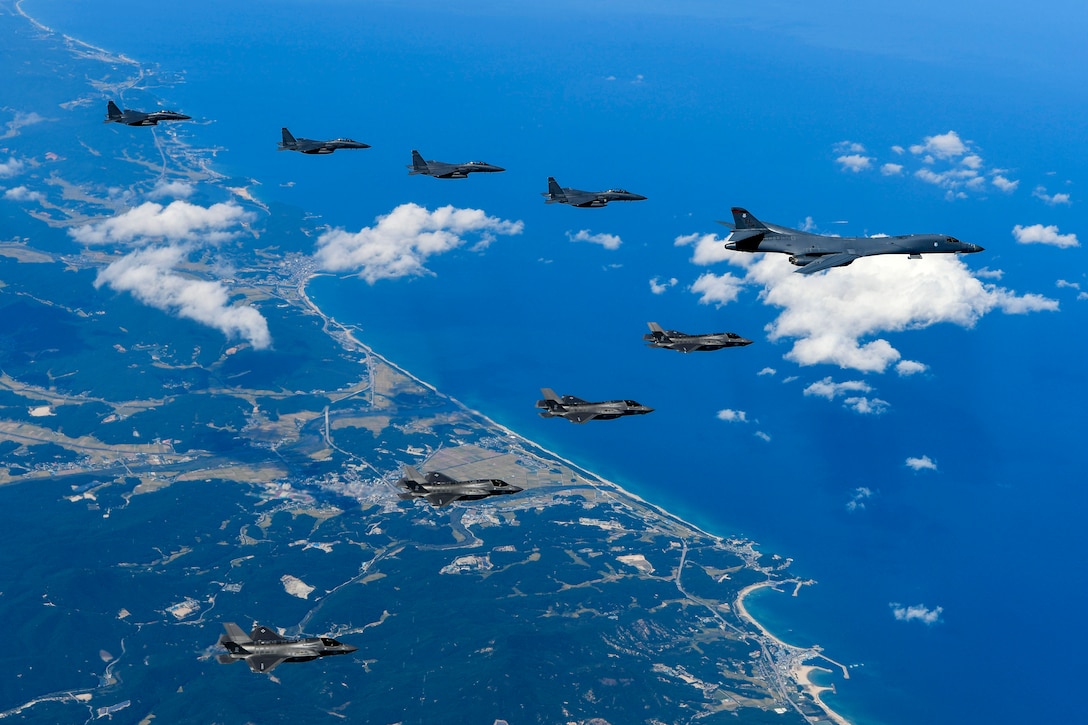 Nine aircraft fly over coastline in a "V" formation.