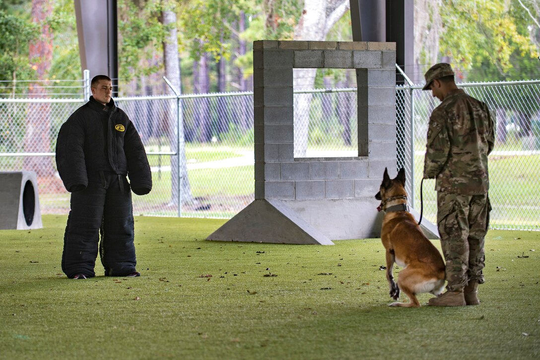 An Airman holds the leash of a dog while a person stands wearing a protective suit.