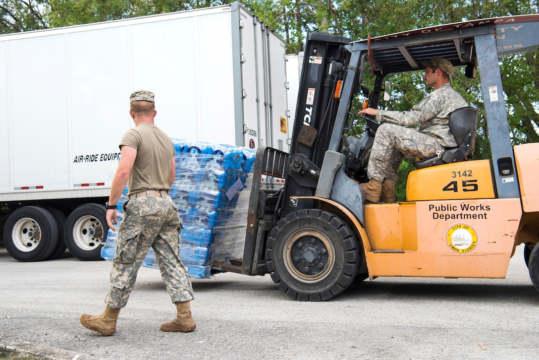 A Guardsman guides a team member operating a forklift transporting pallet of water.