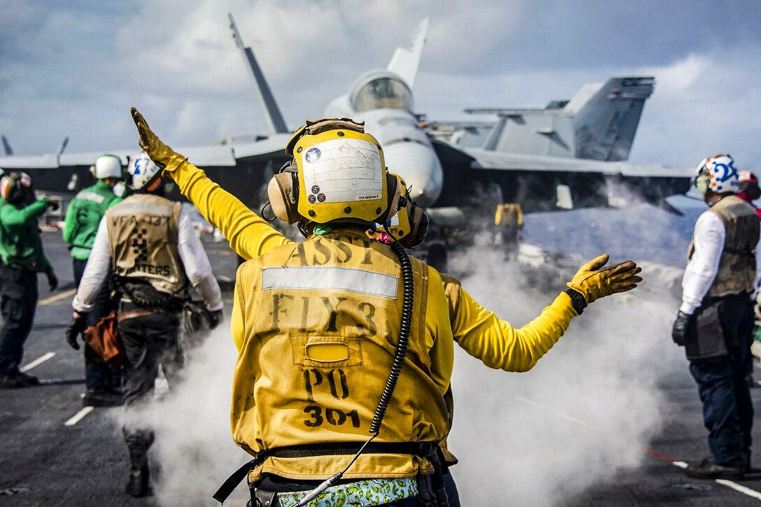 Two sailors use hand signals during flight operations on a deck.