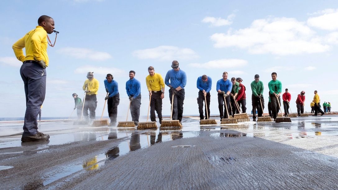 A group of sailors use brushes to scrub the deck of a ship while one blows a whistle.