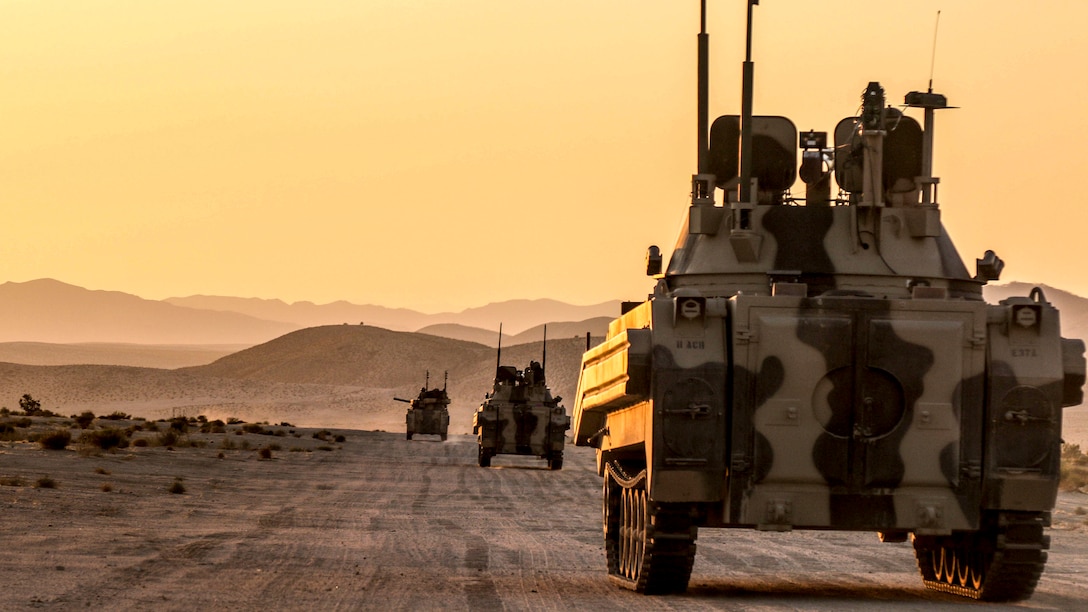 Soldiers drive three military vehicles along a dirt road over a terrain.