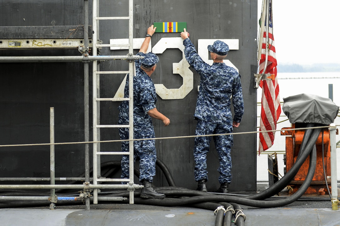 Two sailors affix a green, red and yellow ribbon on an exterior submarine wall above its hull number.