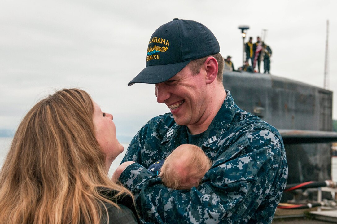 A sailor cradles a baby and smiles at a woman, as a submarine sits docked in the background.