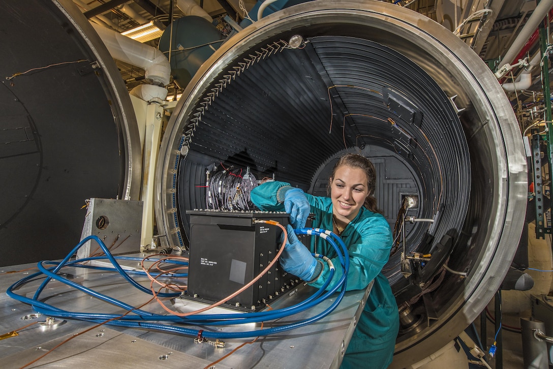 A researcher wearing gloves manipulates wires on a rectangular device in front of a giant metal circular object.