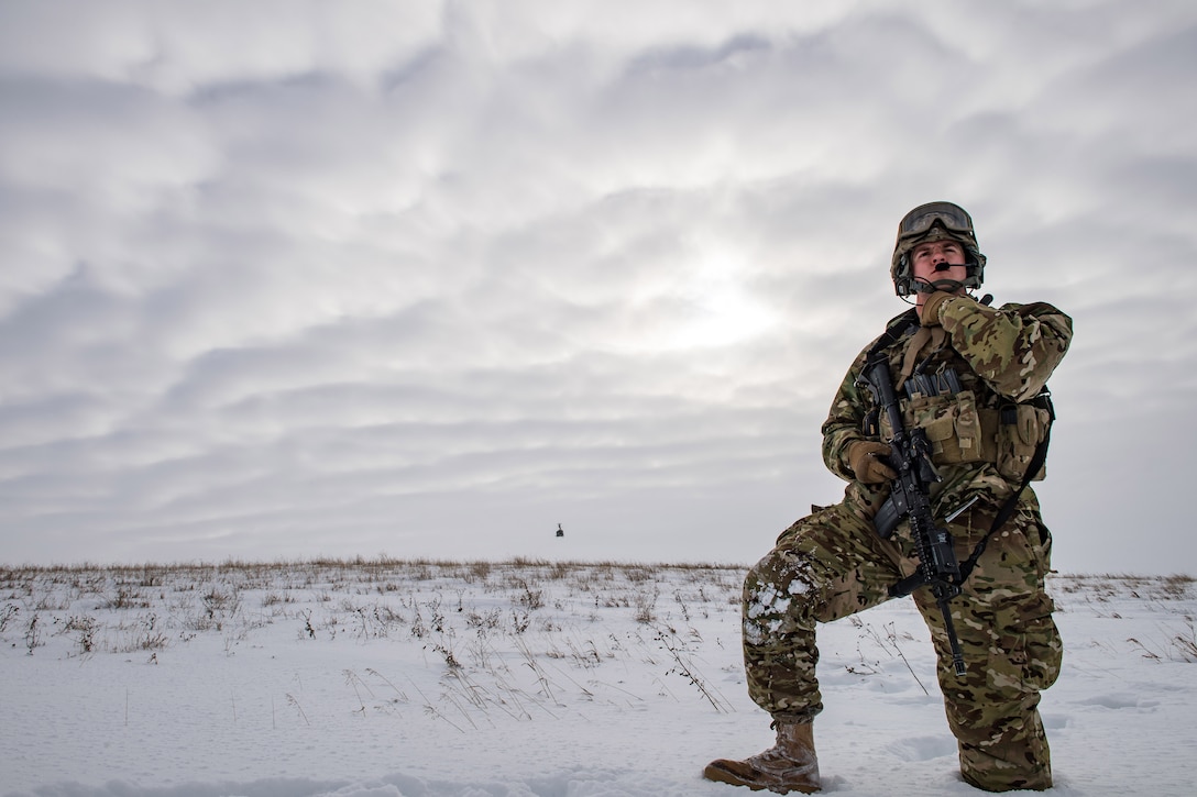 An airman in winter gear stands in a field of snow and looks into the distance.