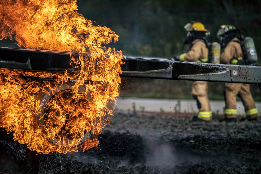 Flames engulf a metal rod as airmen in firefighting garb maneuver in the background.