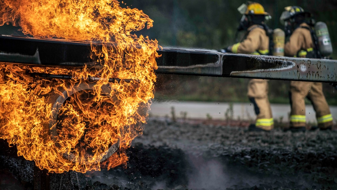 Flames engulf a metal rod as airmen in firefighting garb maneuver in the background.