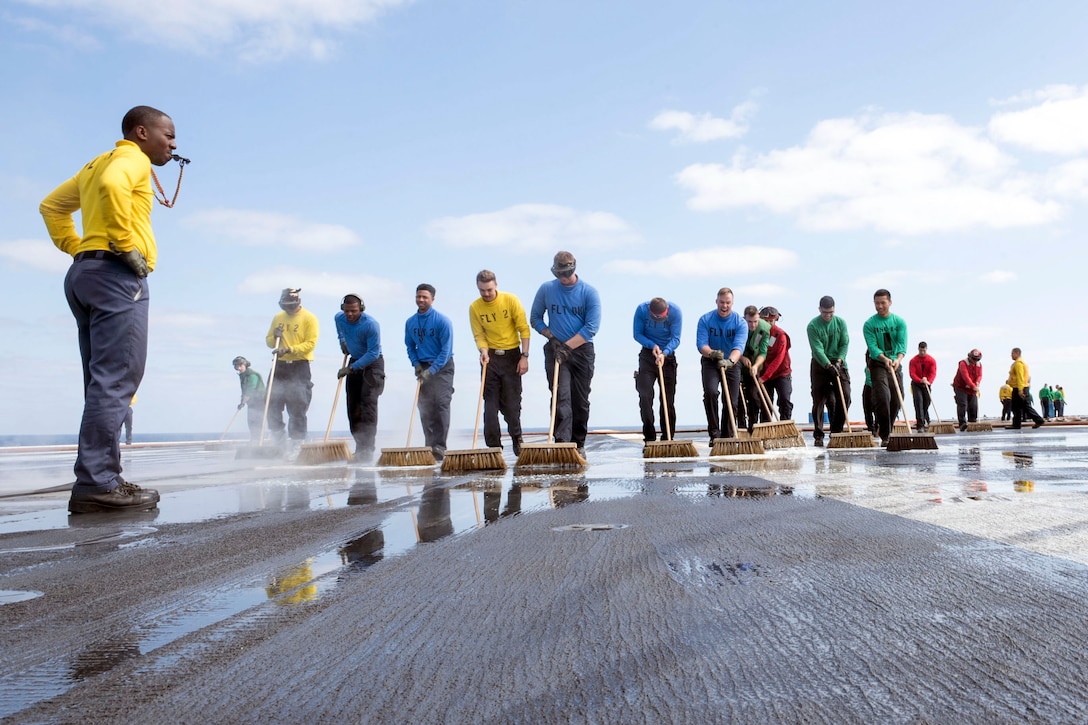 A group of sailors use brushes to scrub the deck of a ship while one blows a whistle.