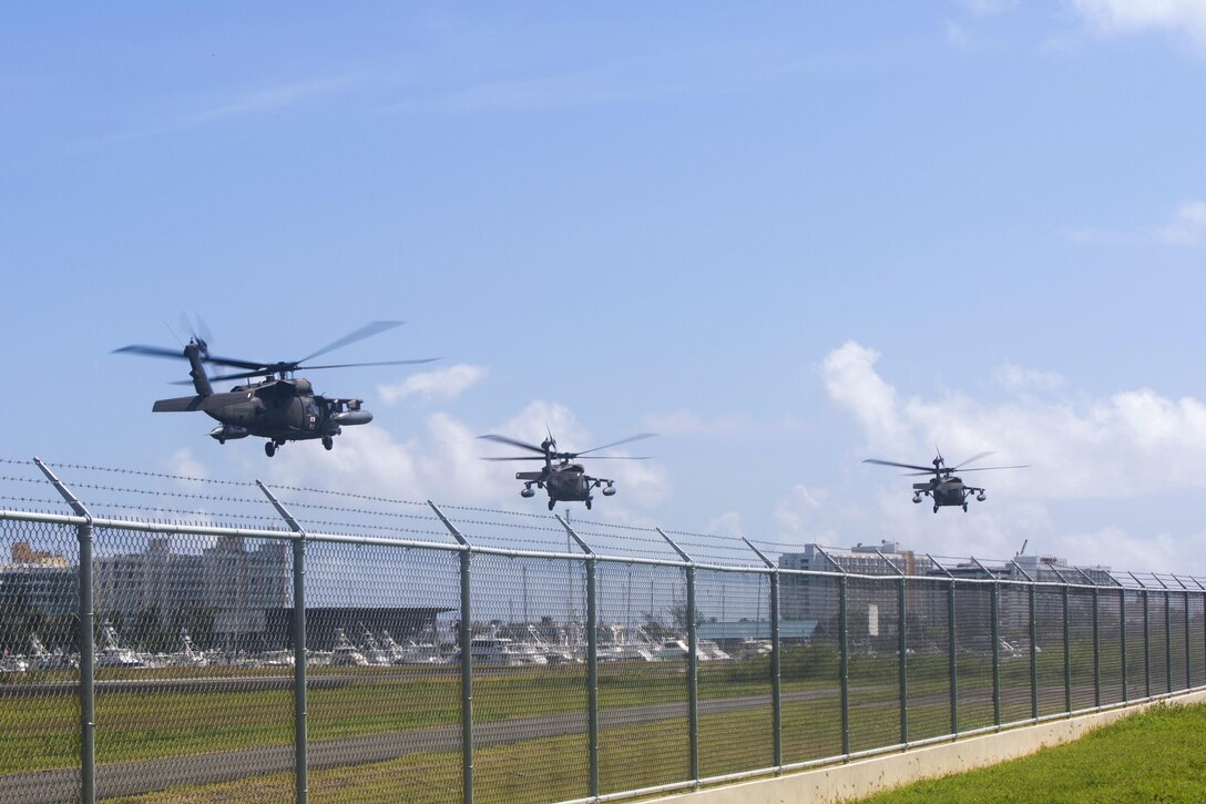 Three helicopters take off from behind a fence.